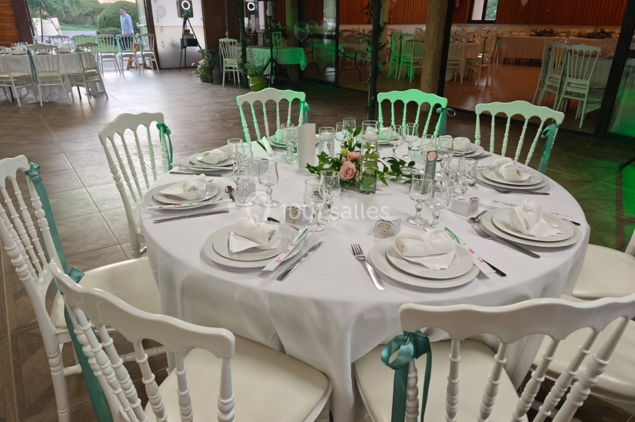 Table ronde élégamment dressée avec nappes blanches, vaisselle raffinée, décoration florale et chaises blanches assorties.