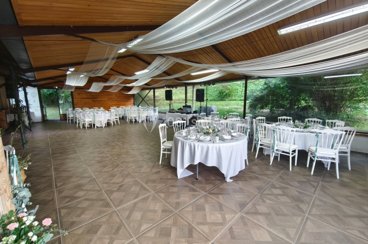 Salle de réception avec tables rondes dressées, chaises blanches et plafond décoré de voilages, ouverte sur un espace…