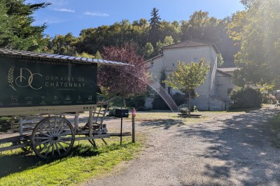Grande maison en pierre entourée de verdure, avec une cour ensoleillée et des arbres en arrière-plan.