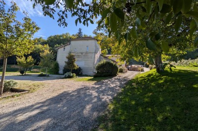 Grande maison en pierre entourée de verdure, avec une cour ensoleillée et des arbres en arrière-plan.