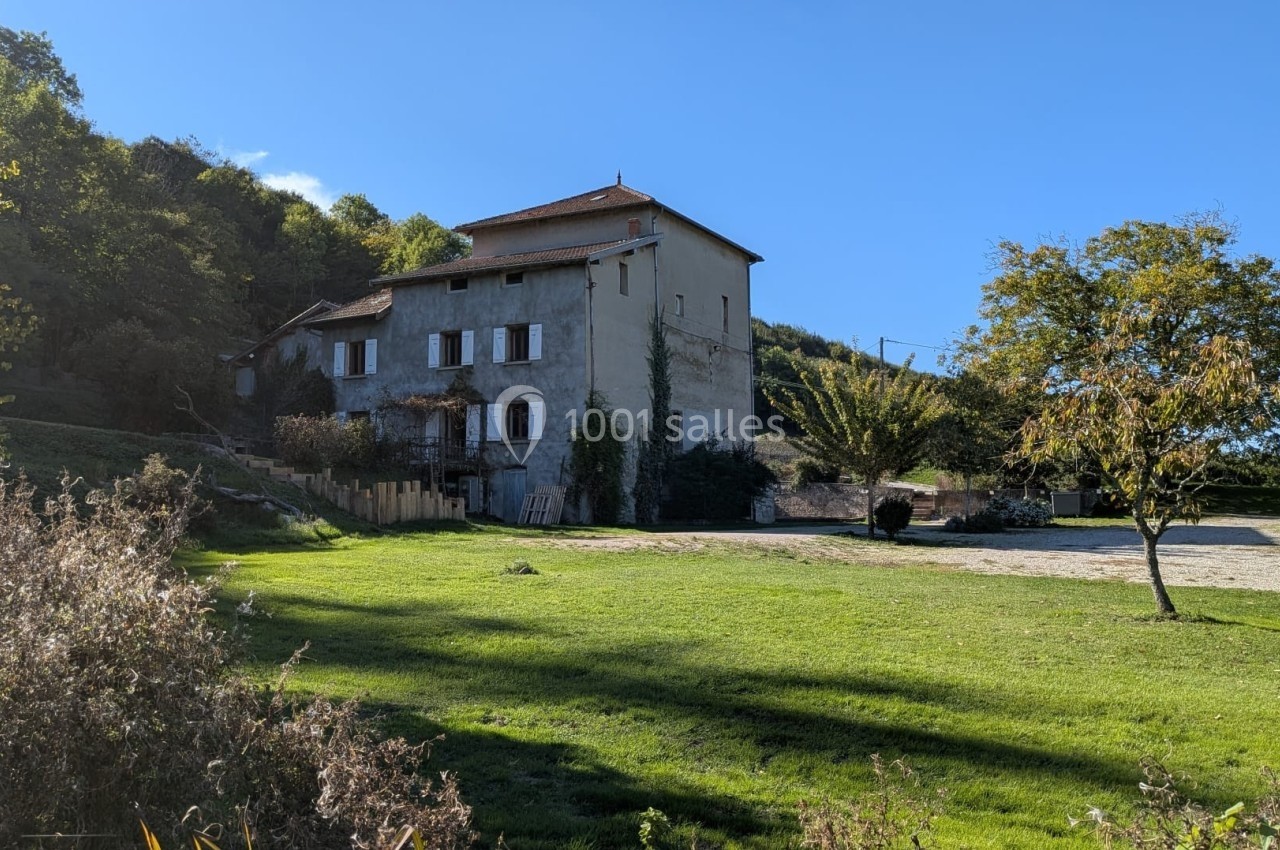Maison en pierre entourée de verdure, avec un arbre au premier plan et une colline boisée en arrière-plan.