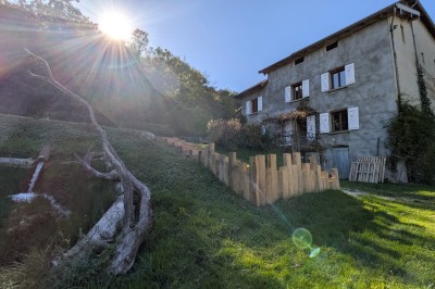 Grande maison en pierre entourée de verdure, avec une cour ensoleillée et des arbres en arrière-plan.