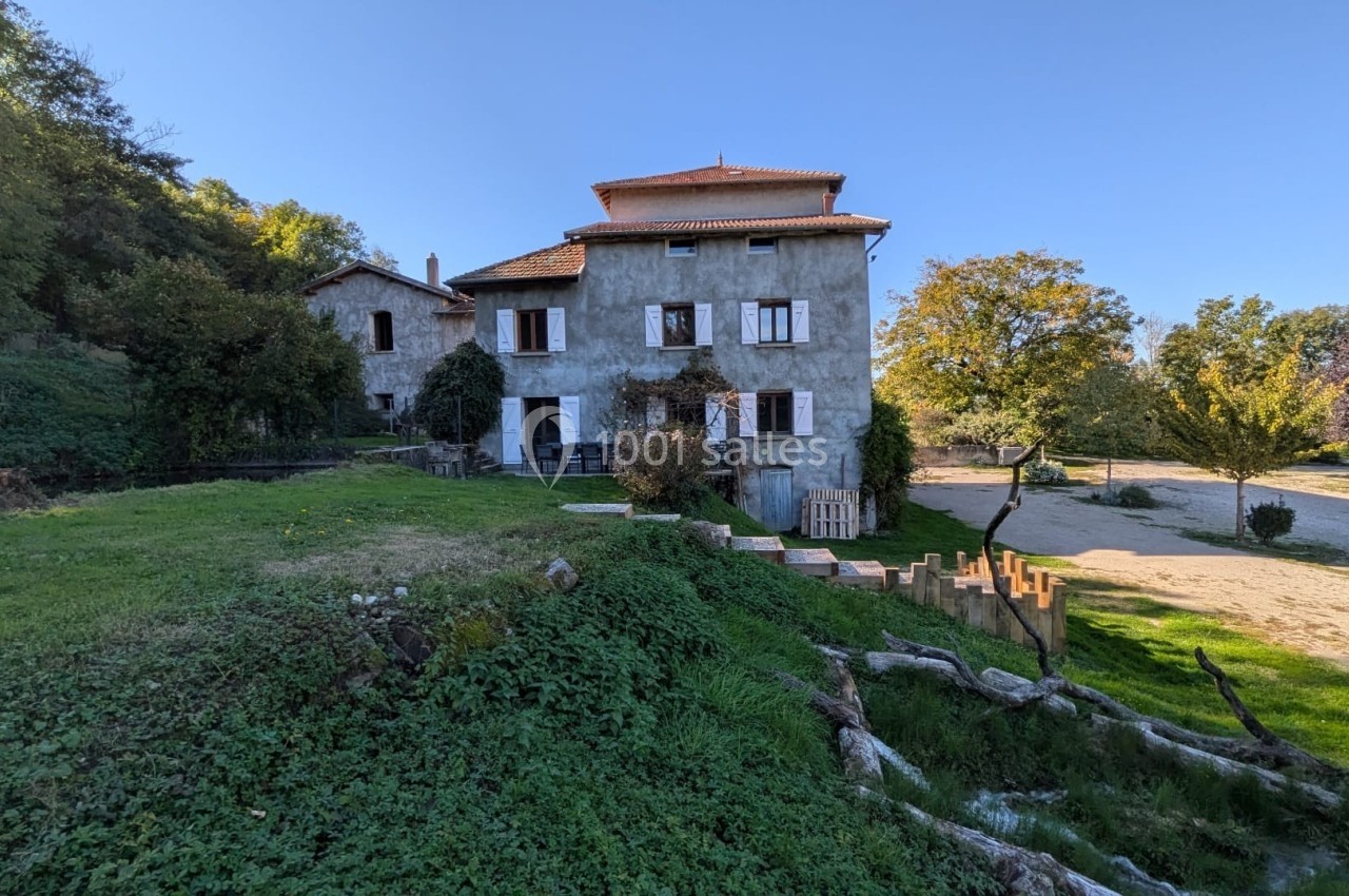 Grande maison en pierre entourée de verdure, avec une cour ensoleillée et des arbres en arrière-plan.