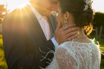 Un couple en tenue de mariage pose dans un champ au coucher du soleil, la mariée tenant un bouquet de fleurs blanches.