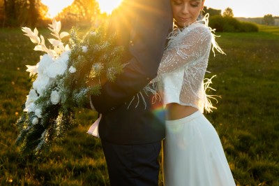 Un couple en tenue de mariage pose dans un champ au coucher du soleil, la mariée tenant un bouquet de fleurs blanches.