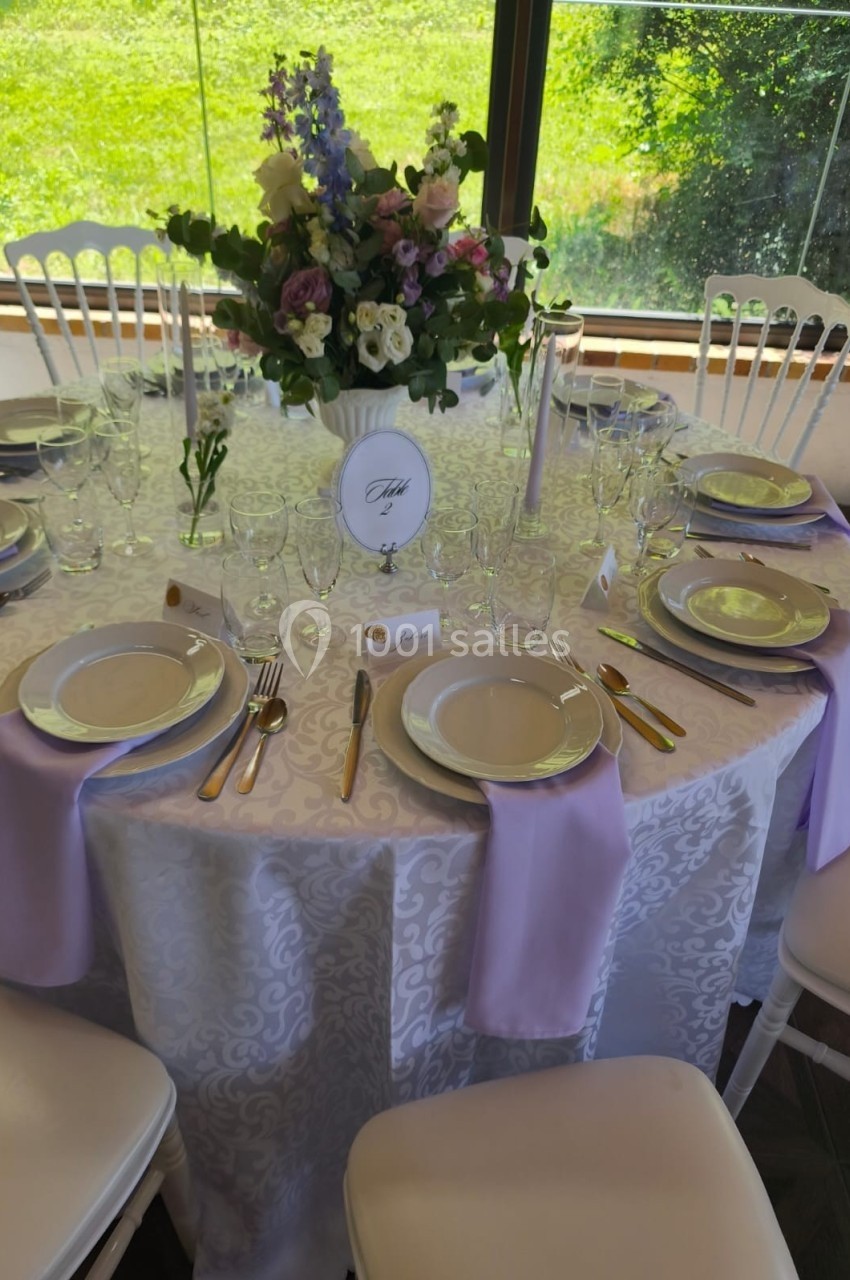 Table ronde élégamment dressée avec nappes blanches, serviettes lavande, vaisselle raffinée et bouquet floral au centre.