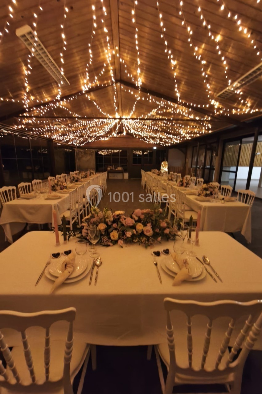 Salle de réception décorée avec des guirlandes lumineuses au plafond, tables dressées avec fleurs et vaisselle élégante.