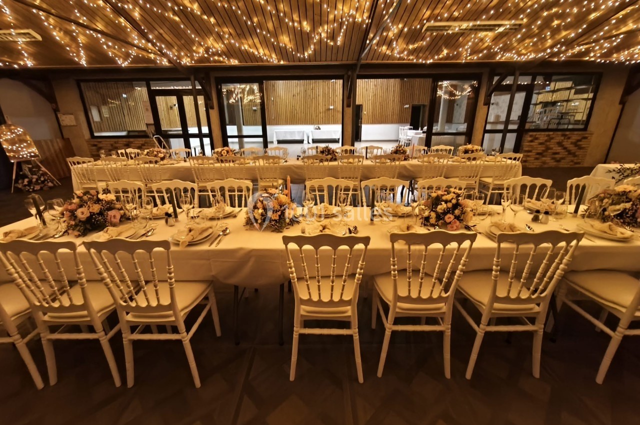 Salle de réception décorée avec des guirlandes lumineuses, tables dressées et chaises blanches alignées.