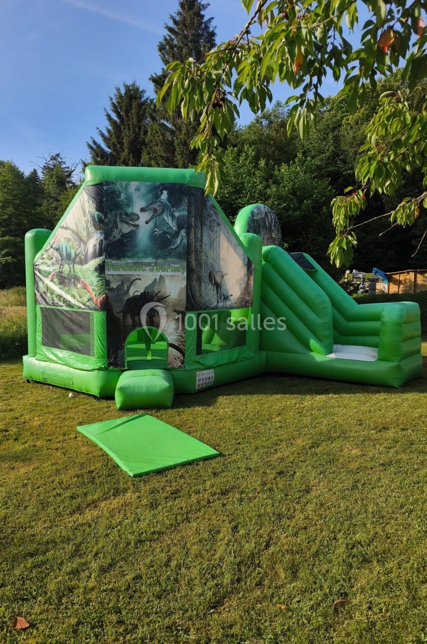 Structure gonflable verte avec toboggan, décorée de motifs de dinosaures, installée sur une pelouse en plein air.