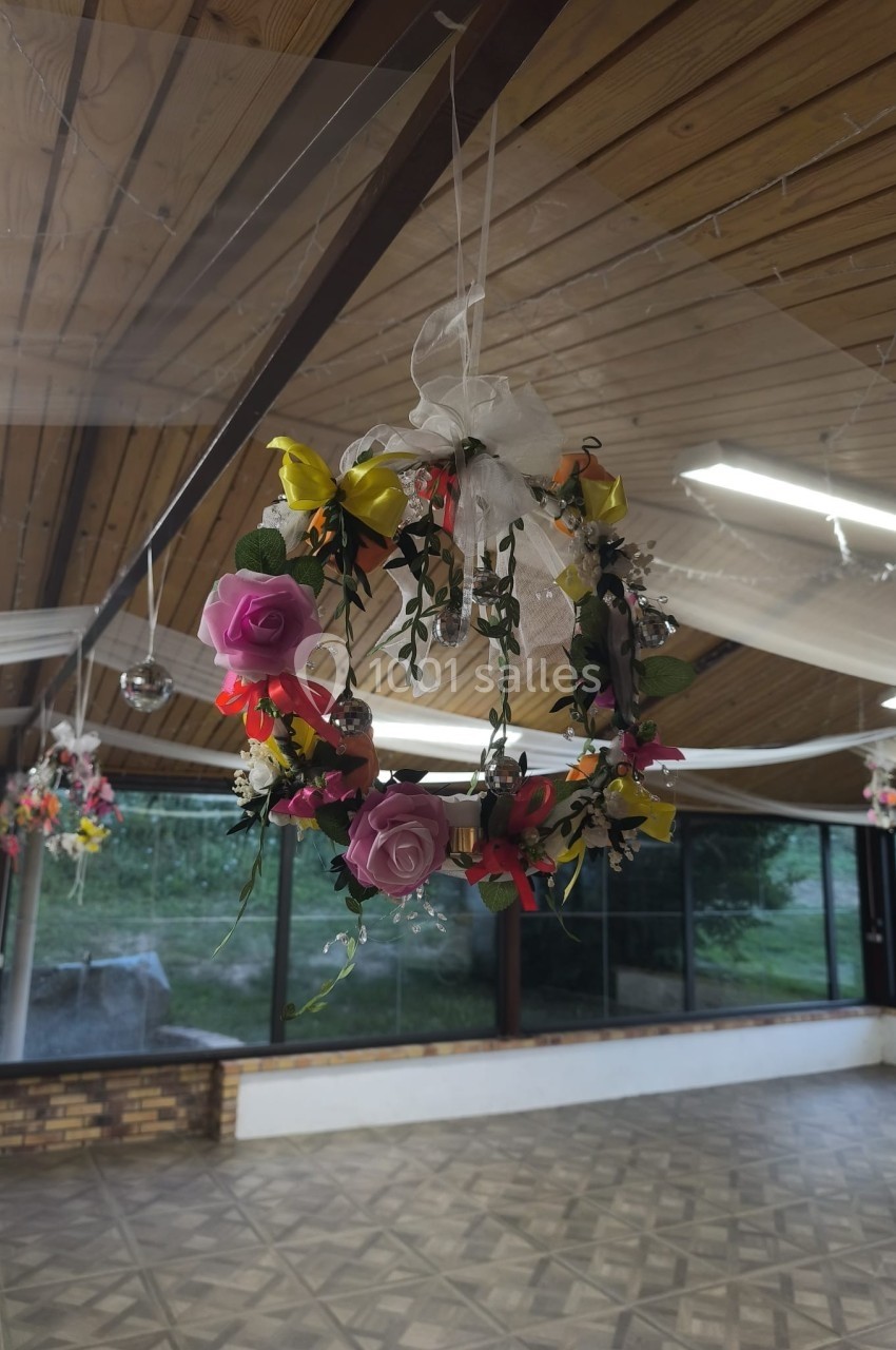 Couronne de fleurs colorées suspendue dans une salle avec plafond en bois et grandes fenêtres donnant sur un jardin.