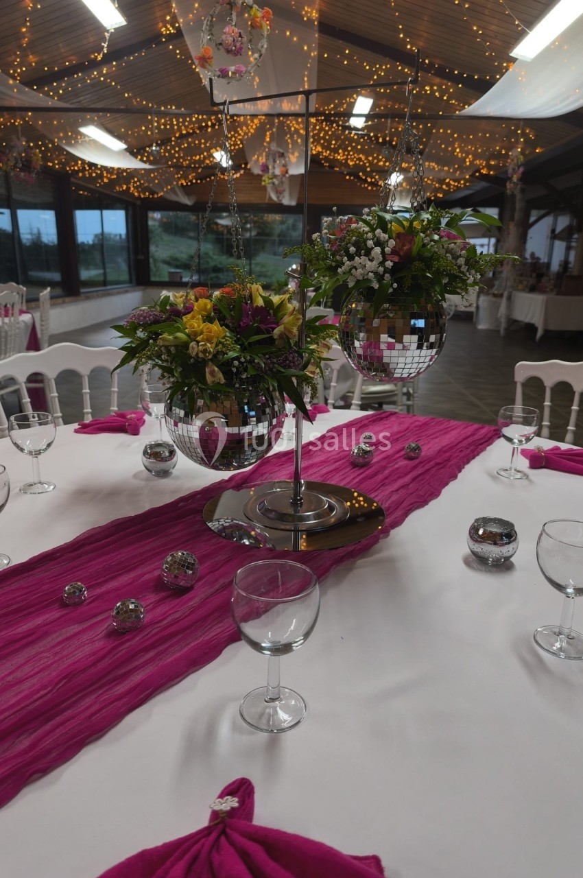 Décoration de table avec nappes blanches, chemin de table fuchsia, fleurs, boules miroir et verres dans une salle éclairée.