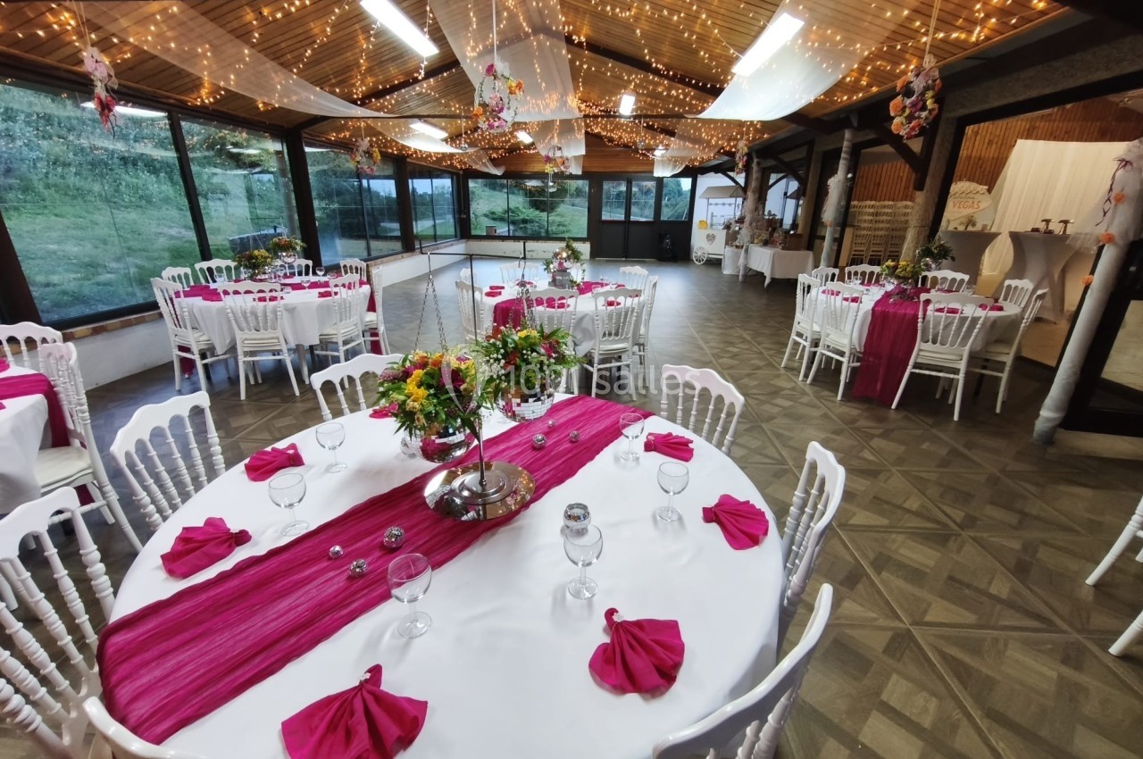 Salle de réception décorée avec des tables rondes, nappes blanches, chemins de table roses et guirlandes lumineuses.