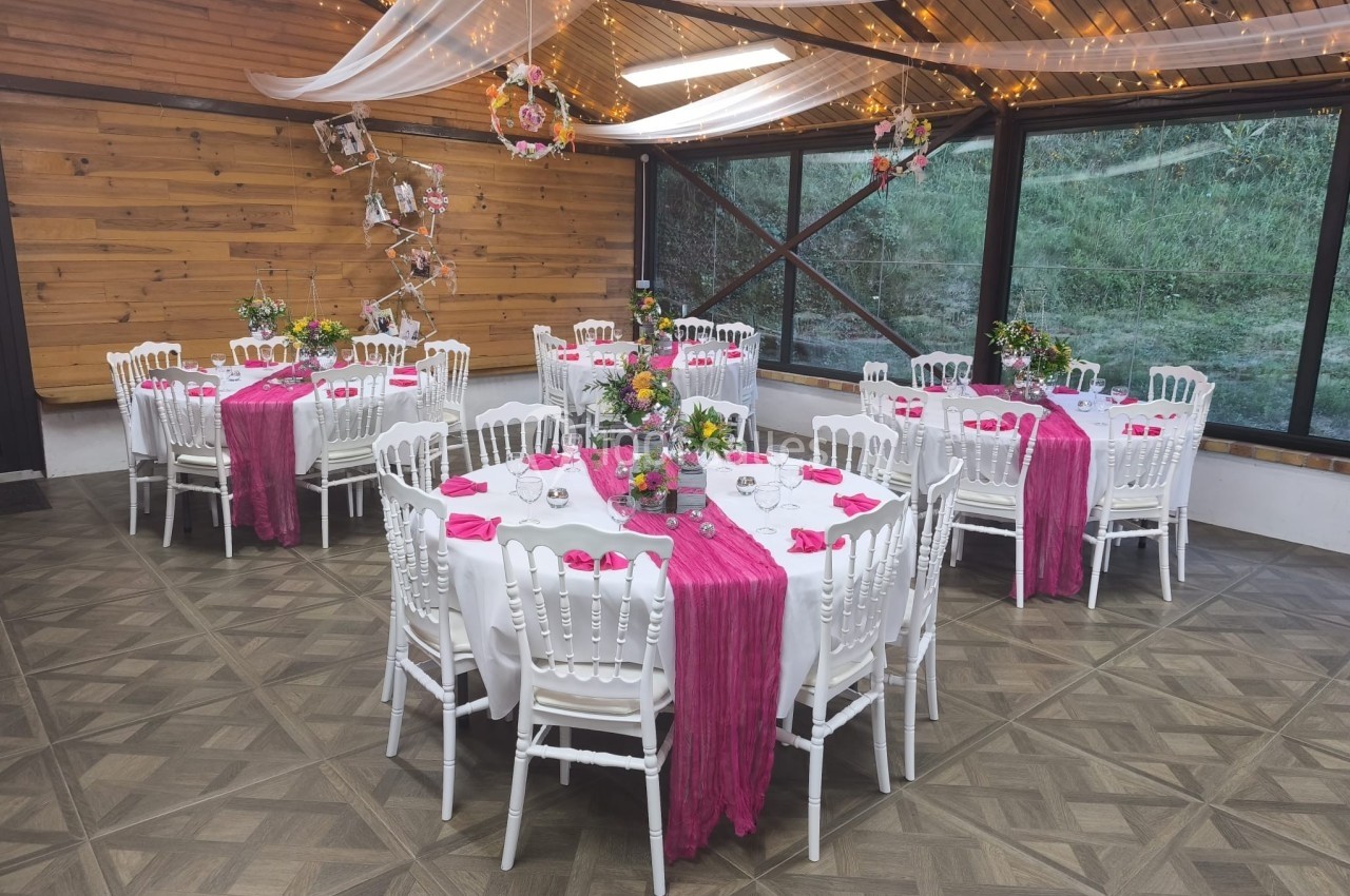 Salle décorée pour un événement avec tables rondes, nappes blanches, chemins de table roses et guirlandes lumineuses.