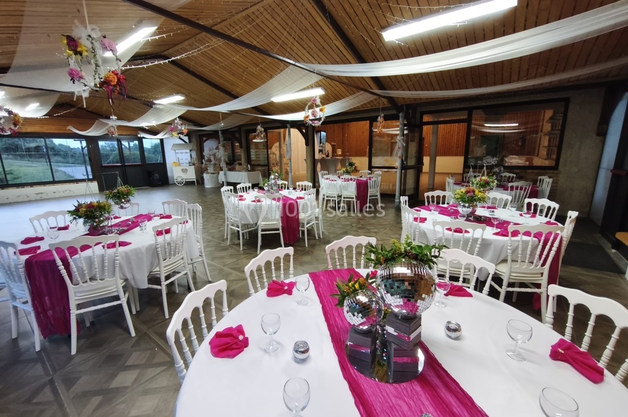 Salle de réception décorée avec des tables rondes, nappes blanches, chemins de table roses et chaises blanches.