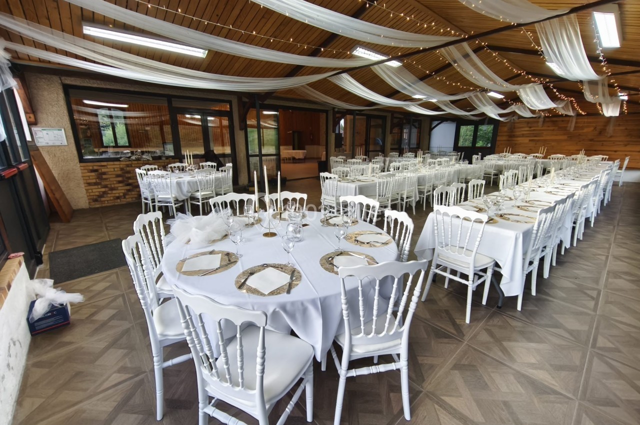 Salle décorée pour un événement, avec tables dressées, nappes blanches, chaises élégantes et guirlandes au plafond.