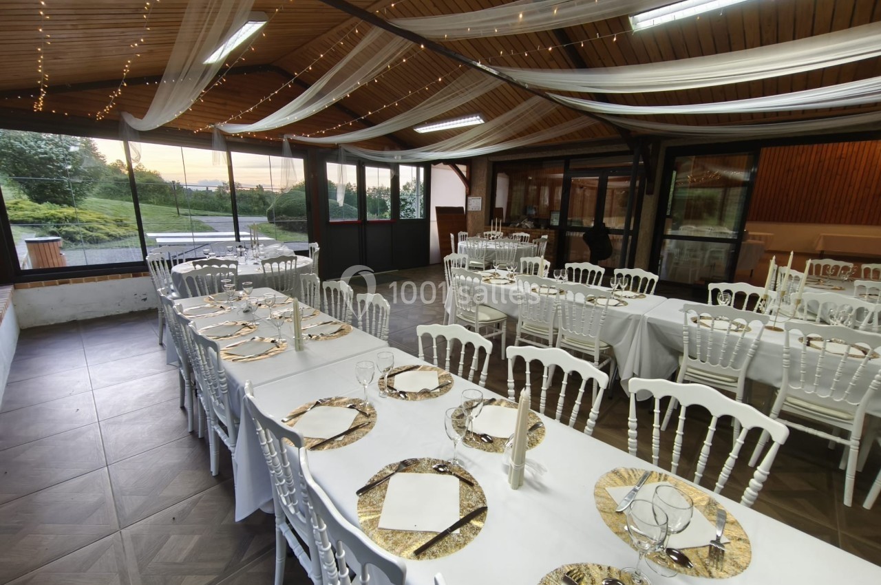 Salle de réception décorée avec des tables dressées, nappes blanches, chaises assorties et voilages suspendus.