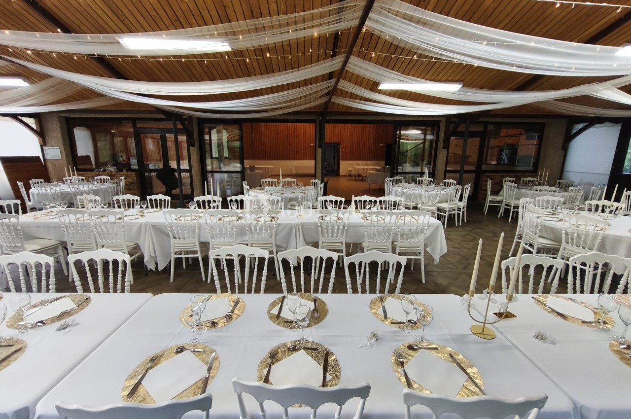 Salle de réception décorée avec des tables dressées, nappes blanches, chaises blanches et chemins de table dorés.
