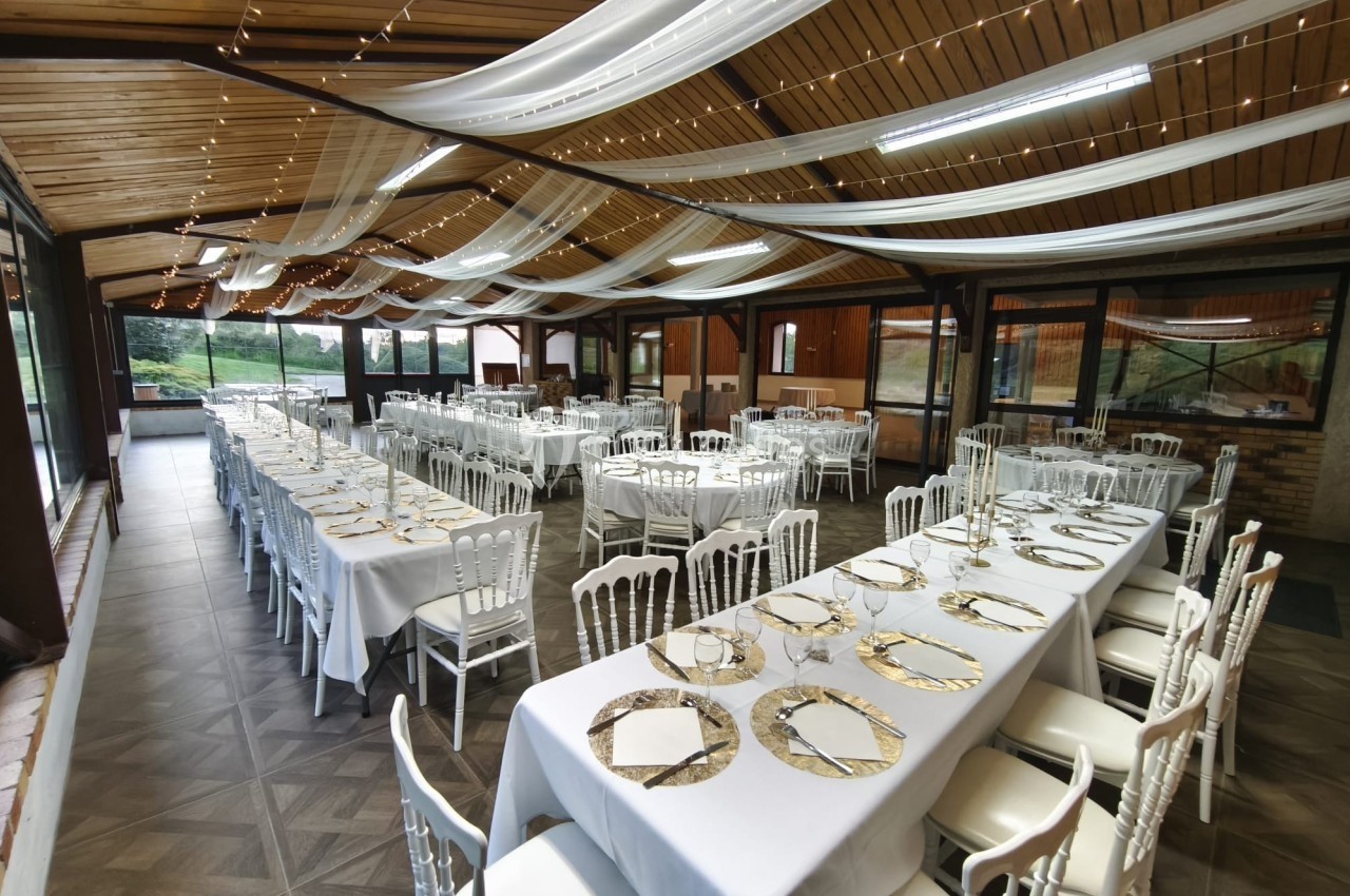 Salle de réception décorée avec des tables alignées, nappes blanches, chaises élégantes et guirlandes lumineuses au plafond.