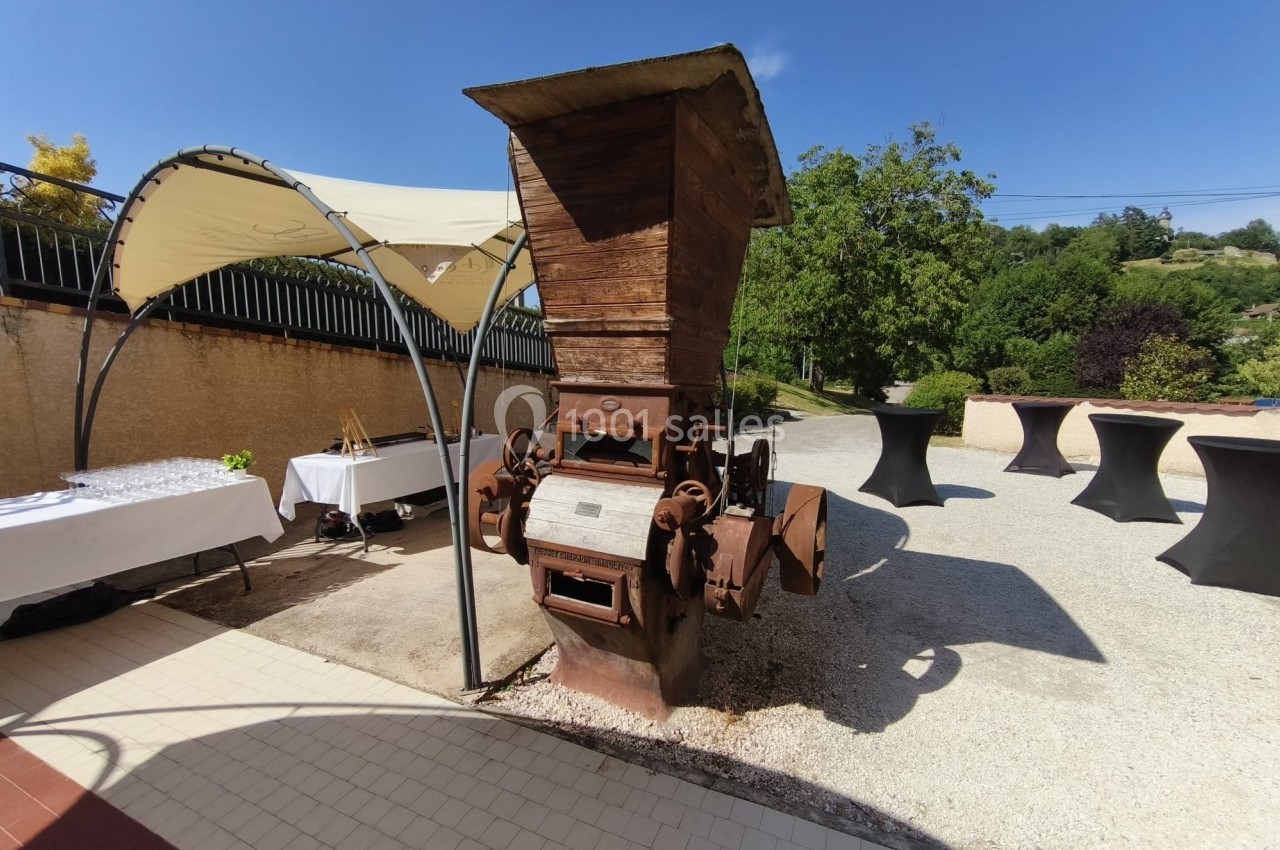 Ancienne machine agricole en métal rouillé exposée en extérieur, entourée de tables et d'arbres sous un ciel bleu.