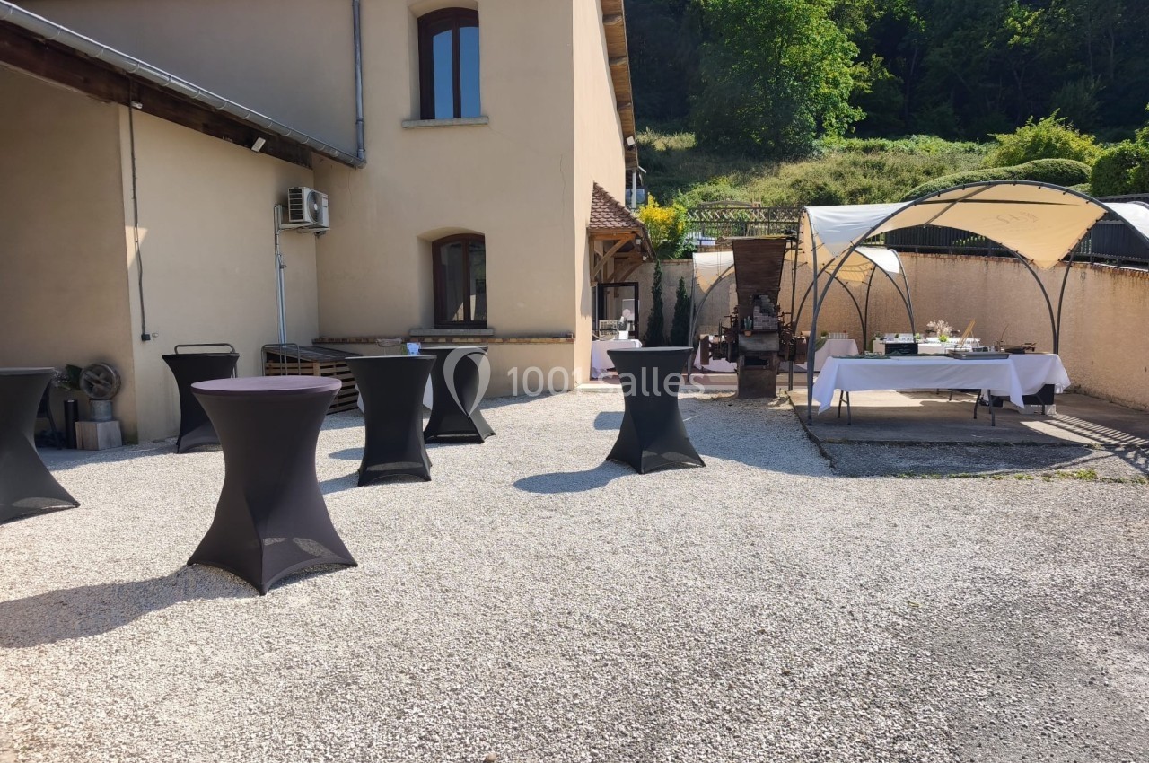 Cour extérieure avec tables hautes noires et buffet sous une tonnelle, entourée de bâtiments et de verdure.