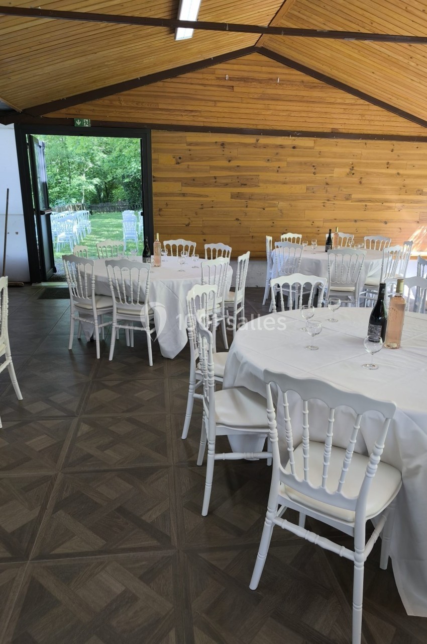 Salle de réception avec tables rondes dressées, nappes blanches et chaises blanches, vue sur un espace extérieur verdoyant.