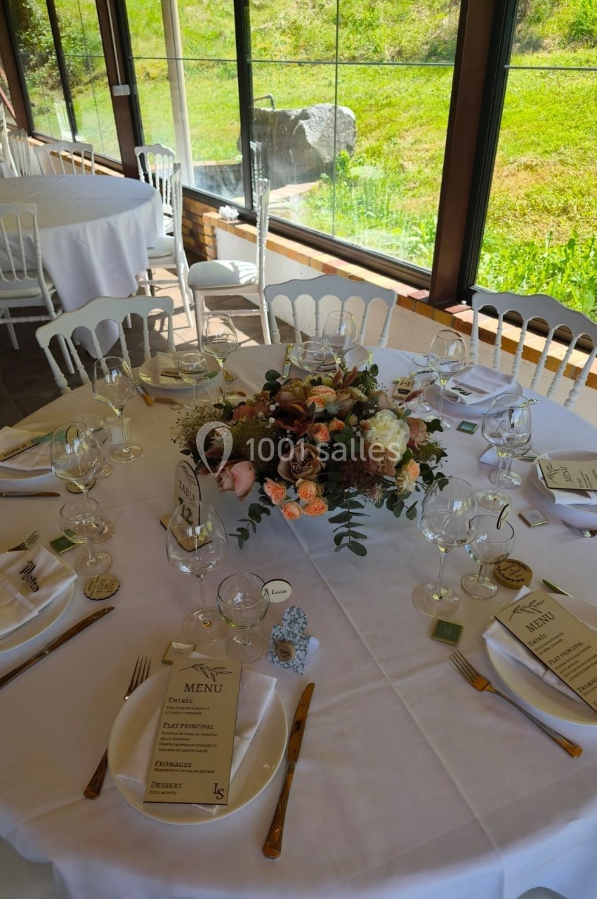 Table de réception élégamment dressée avec nappes blanches, menus, fleurs et couverts dorés, près d'une baie vitrée.