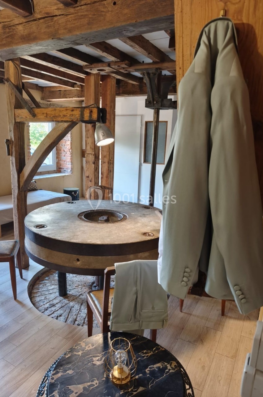 Deux vestes beiges suspendues dans une pièce rustique avec une table ronde en pierre et des poutres en bois apparentes.