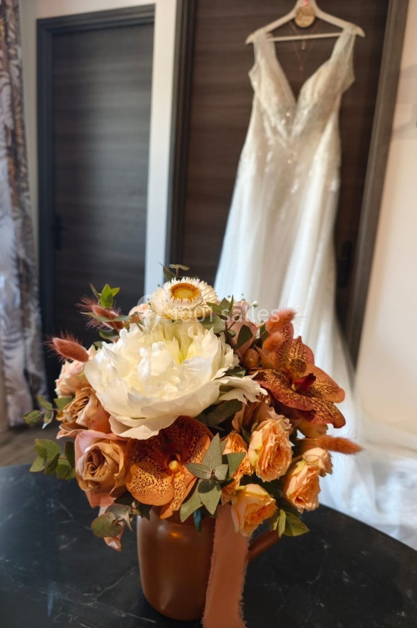 Bouquet de fleurs aux tons chauds posé sur une table, avec une robe de mariée suspendue en arrière-plan.