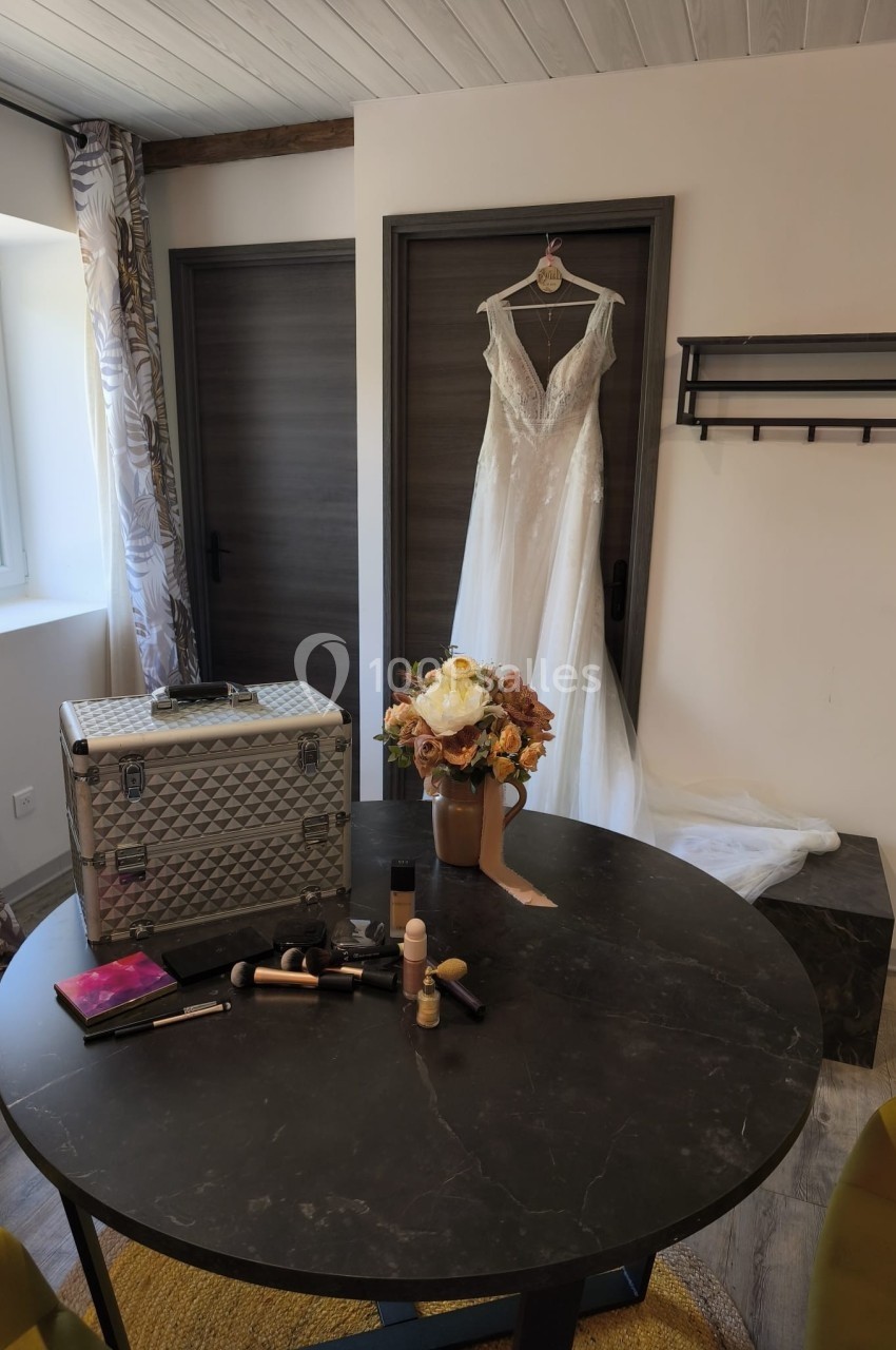 Robe de mariée suspendue à une porte, bouquet de fleurs et maquillage disposés sur une table ronde.