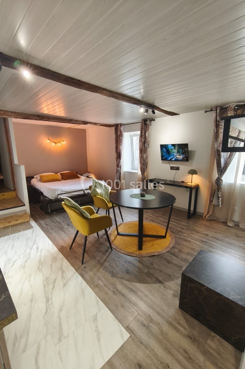 Studio lumineux avec lit double, coin salon, table ronde, chaises jaunes, télévision murale et parquet clair.