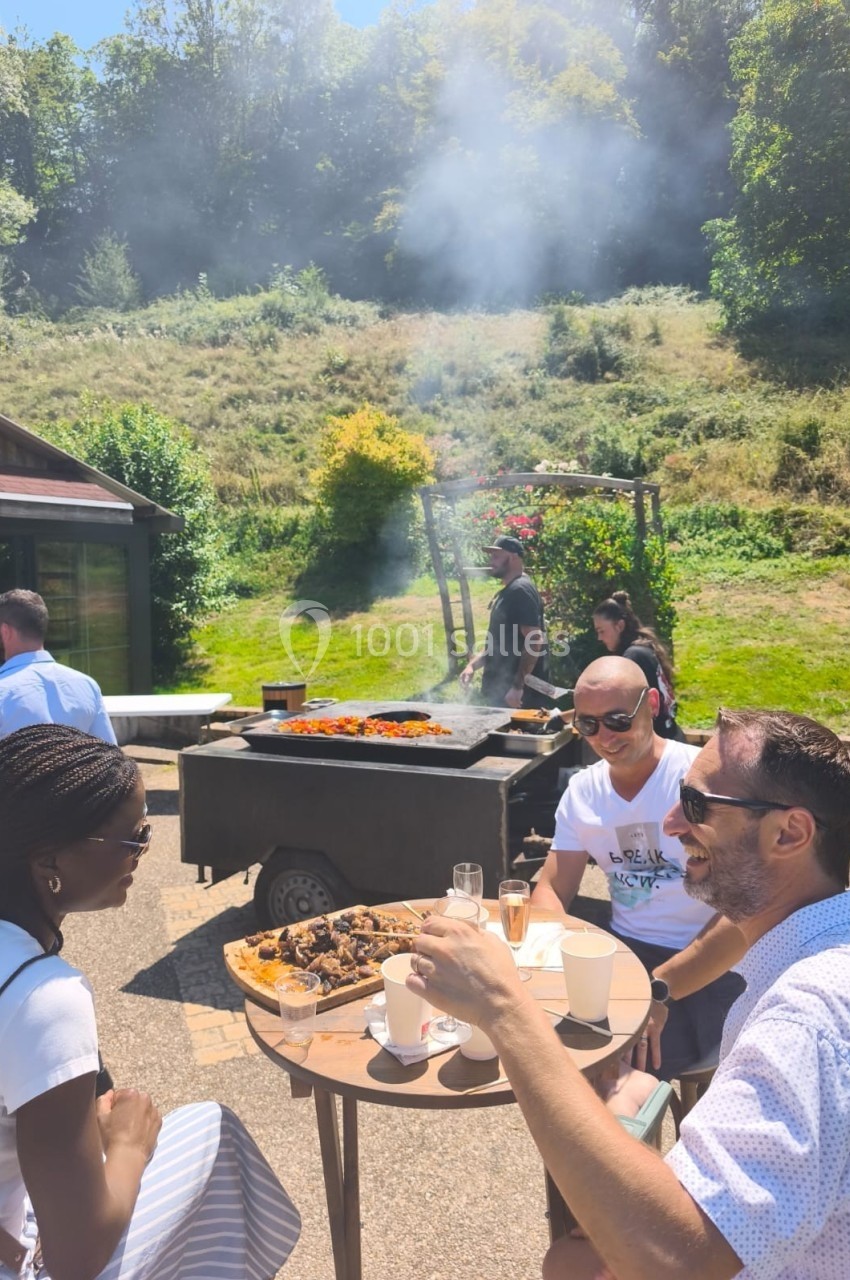 Des personnes discutent autour d'une table en extérieur près d'un barbecue en pleine cuisson, sous un ciel ensoleillé.
