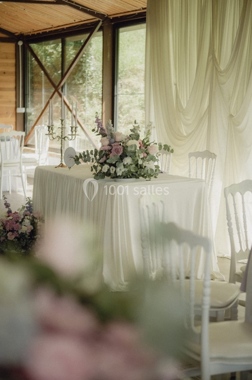 Table décorée avec une nappe blanche, un bouquet floral et des chandeliers, dans une salle lumineuse avec rideaux et baies…