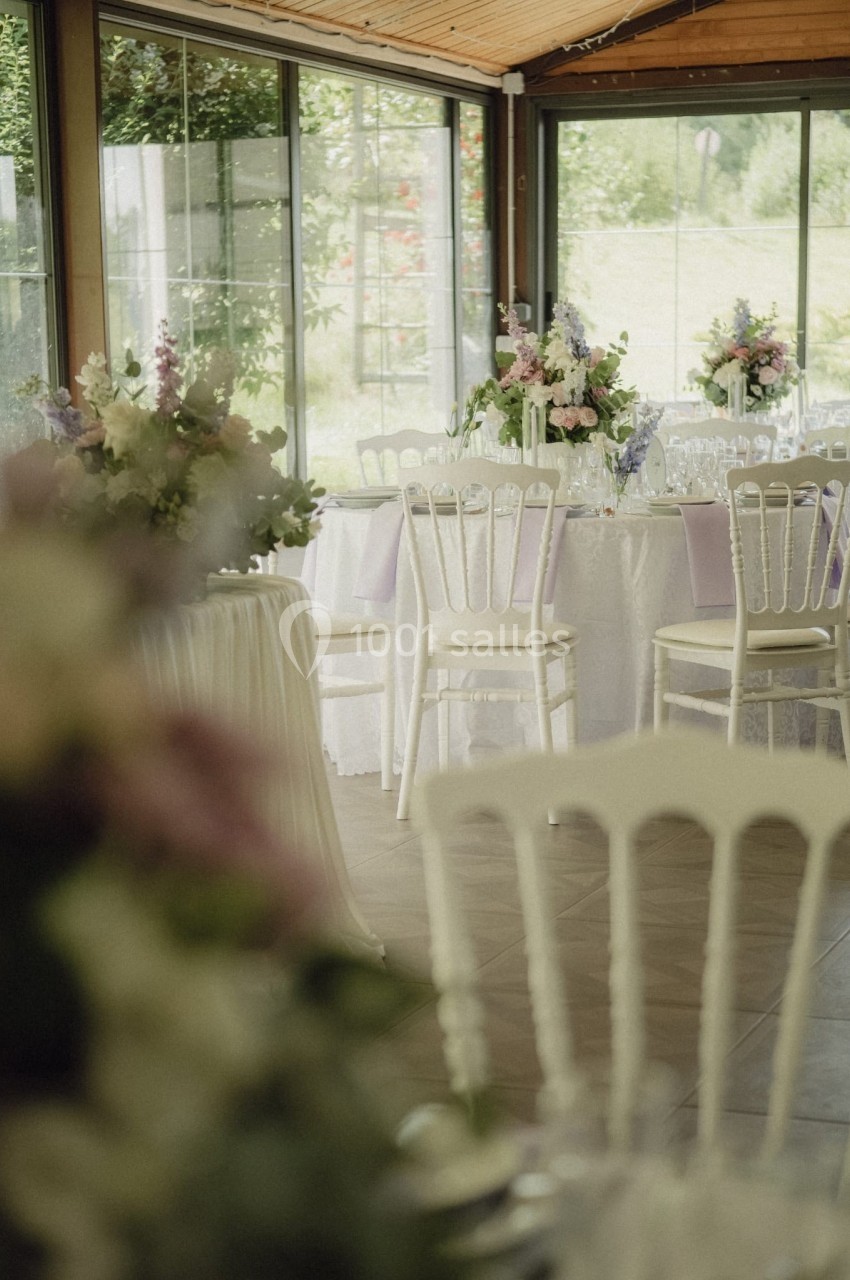 Salle lumineuse décorée pour un événement, avec des tables rondes, nappes blanches et bouquets de fleurs.