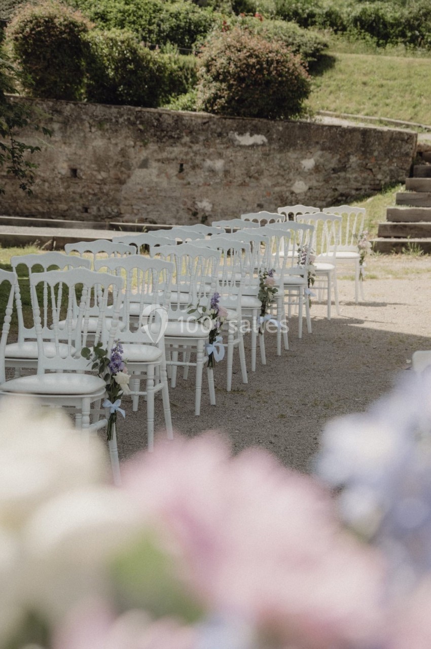 Chaises blanches alignées en extérieur, décorées de fleurs, sur un chemin gravillonné près d'un mur en pierre.