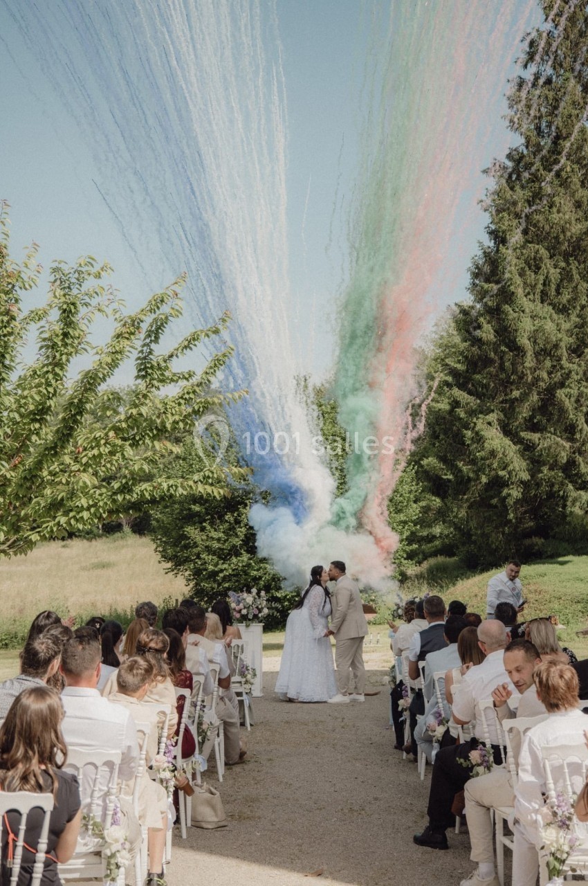 Un couple s'embrasse lors d'une cérémonie de mariage en plein air, avec des fumées colorées en arrière-plan.