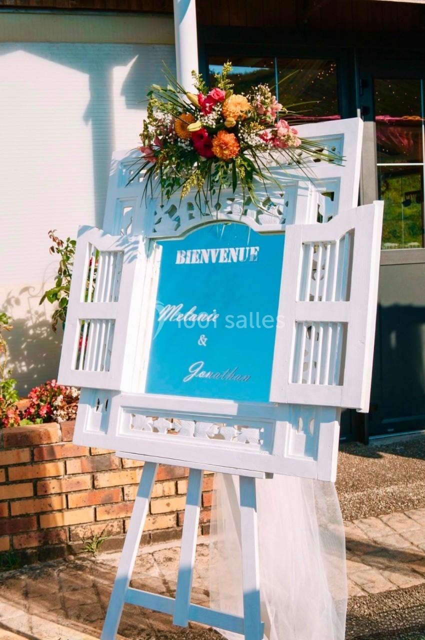 Panneau de bienvenue décoré de fleurs colorées, posé sur un chevalet blanc devant un bâtiment.
