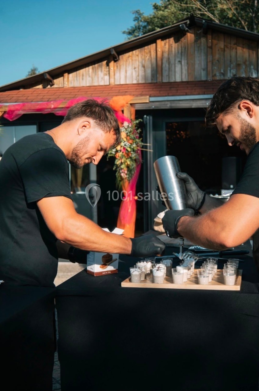 Deux hommes préparent des boissons dans des gobelets sur une table en extérieur, devant un bâtiment en bois.