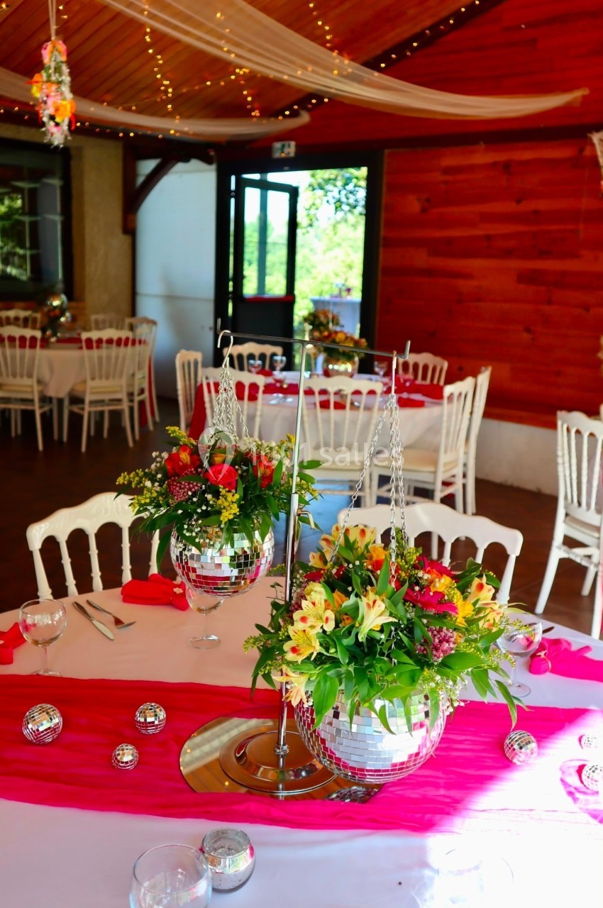 Salle de réception décorée avec des tables ornées de nappes roses, fleurs colorées et boules à facettes suspendues.
