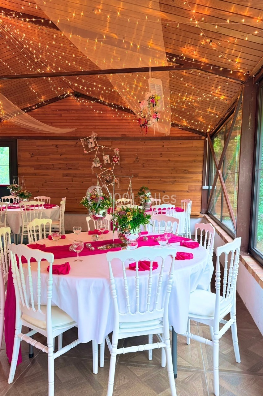 Salle de réception en bois décorée avec des guirlandes lumineuses, tables rondes dressées avec nappes blanches et touches de…