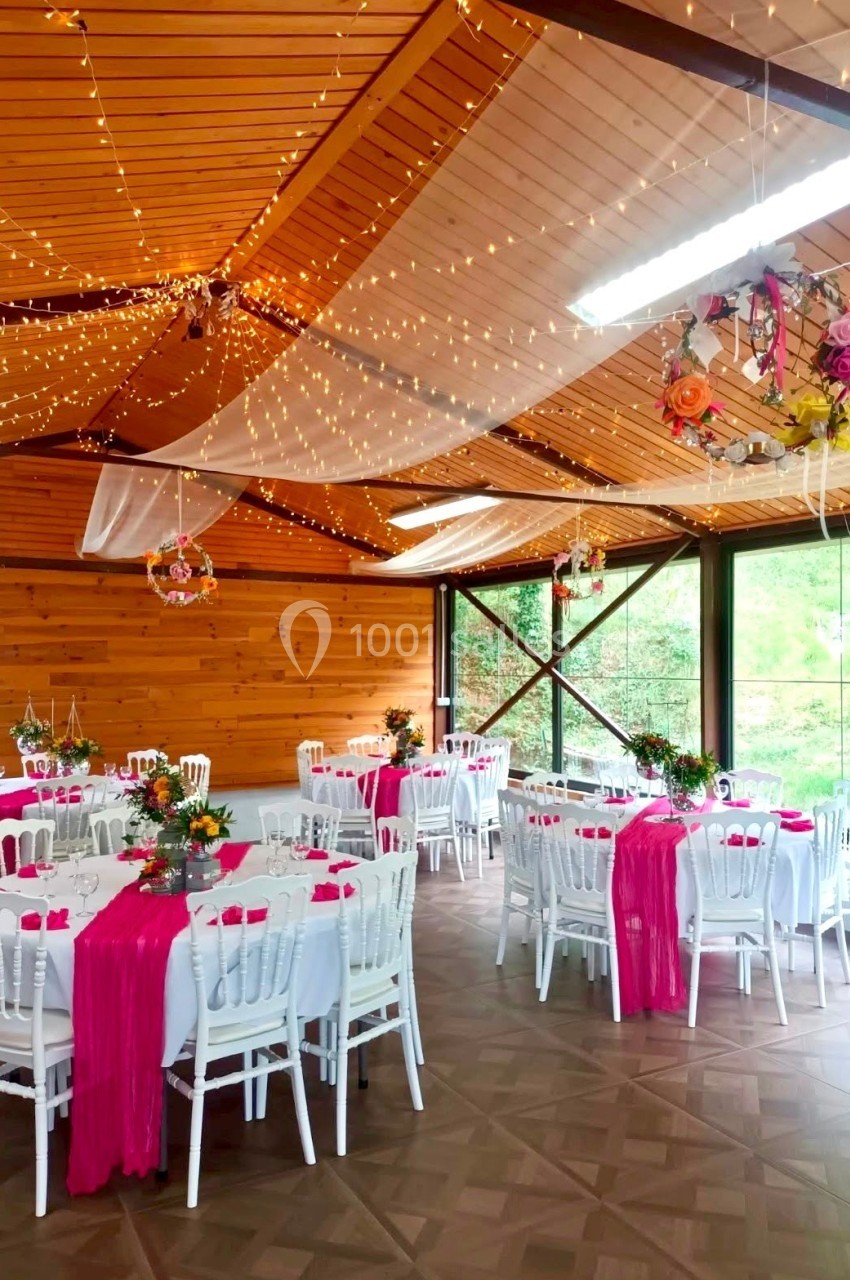 Salle décorée pour un événement, avec des tables rondes ornées de nappes blanches et chemins de table roses, éclairée par…