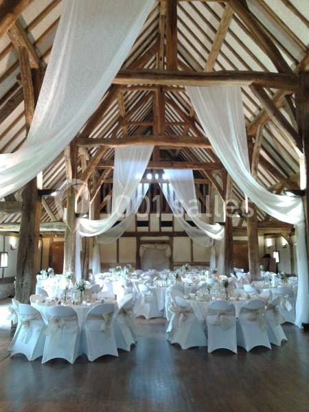 Salle de réception en bois avec poutres apparentes, tables rondes dressées et drapés blancs suspendus.