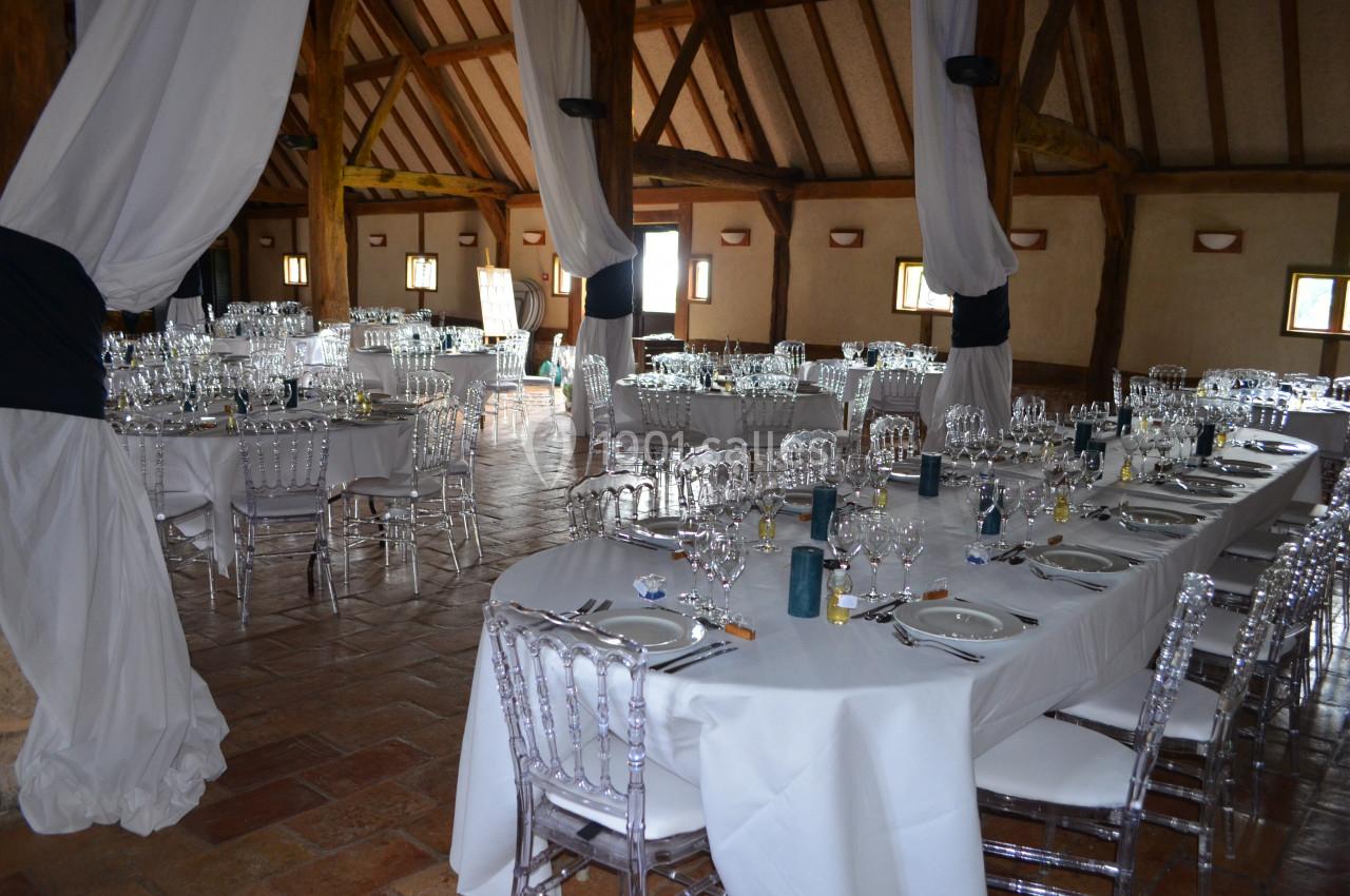 Salle de réception décorée avec des tables dressées, nappes blanches, chaises transparentes et poutres apparentes.