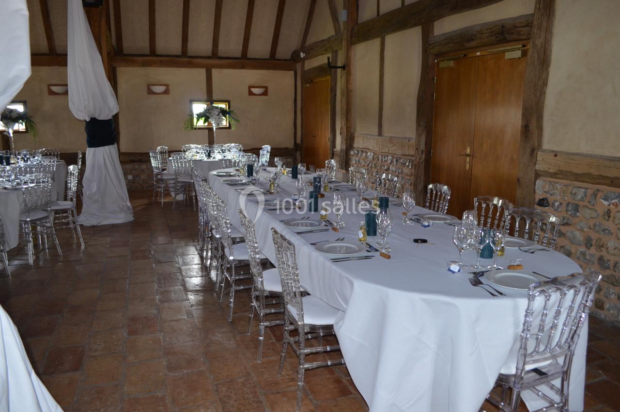 Salle rustique avec tables dressées de nappes blanches, chaises transparentes et vaisselle prête pour un repas.