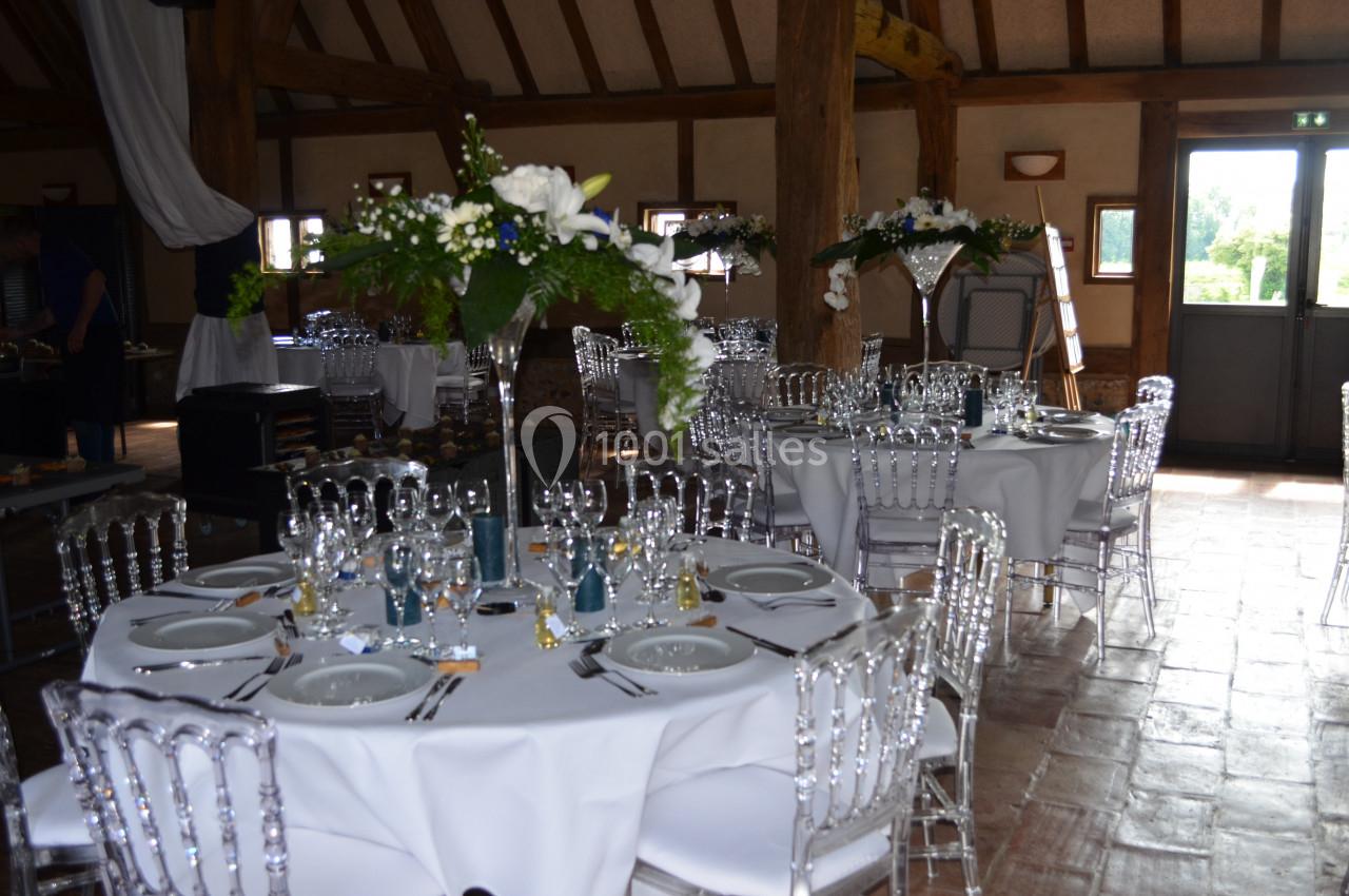 Salle de réception avec tables rondes dressées, chaises transparentes et décorations florales blanches et vertes.
