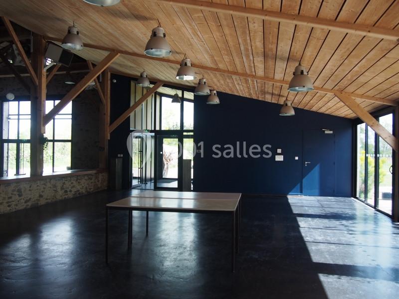 Salle lumineuse avec murs bleus, plafond en bois, grandes fenêtres et une table au centre sur un sol sombre.