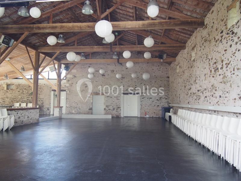 Salle spacieuse avec murs en pierre, plafond en bois, lampes suspendues et chaises blanches alignées le long des murs.