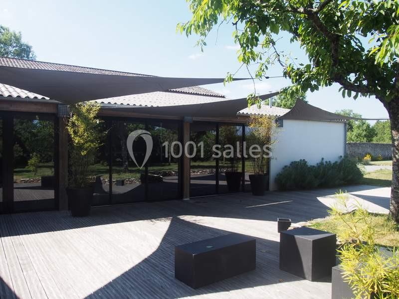 Terrasse en bois avec mobilier moderne, ombragée par des voiles tendues, entourée de végétation et d'un bâtiment vitré.