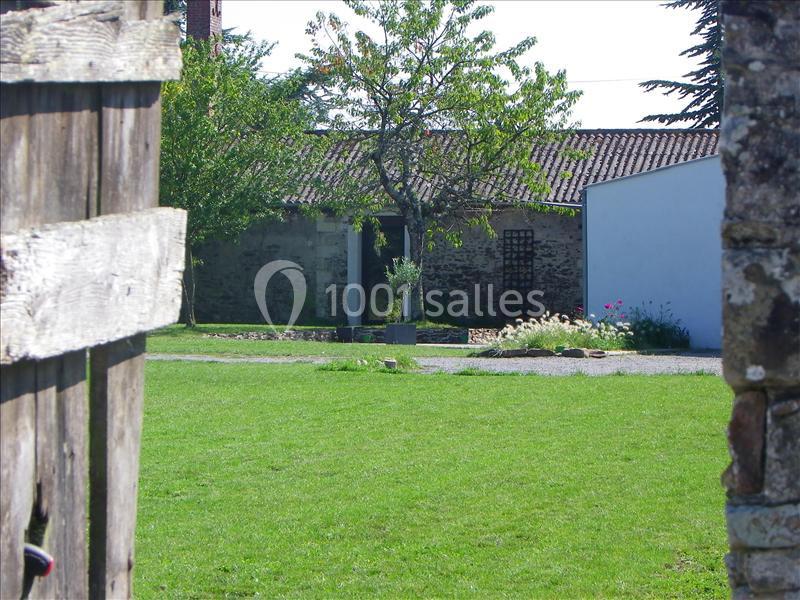 Vue d'une cour verdoyante avec pelouse, arbres et bâtiments en pierre sous un ciel dégagé, encadrée par une clôture en bois.