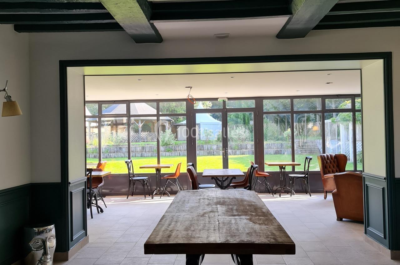 Vue intérieure d'une pièce lumineuse avec une grande table en bois, des poutres apparentes et des baies vitrées donnant sur…