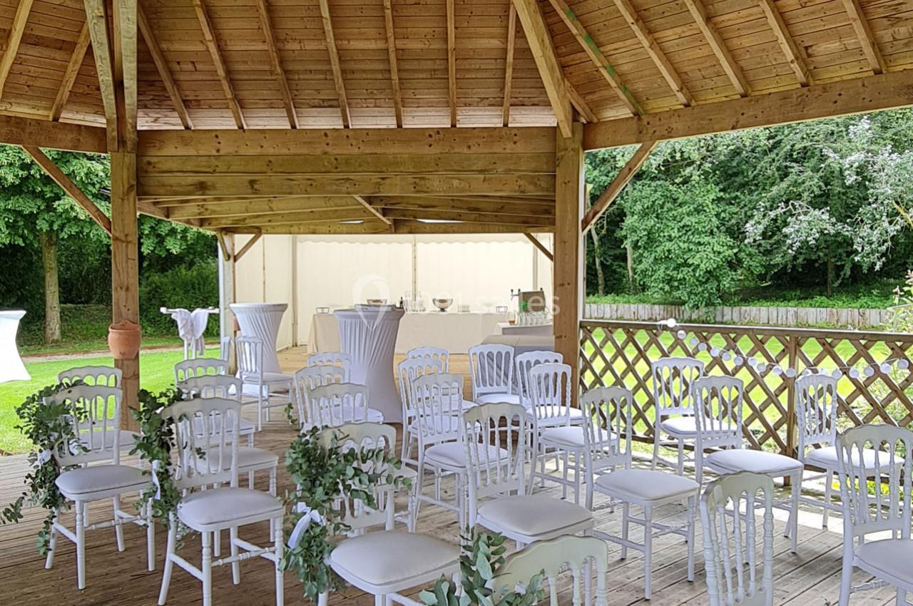 Chaises blanches disposées sous une pergola en bois, entourée de verdure, avec un espace de réception en arrière-plan.